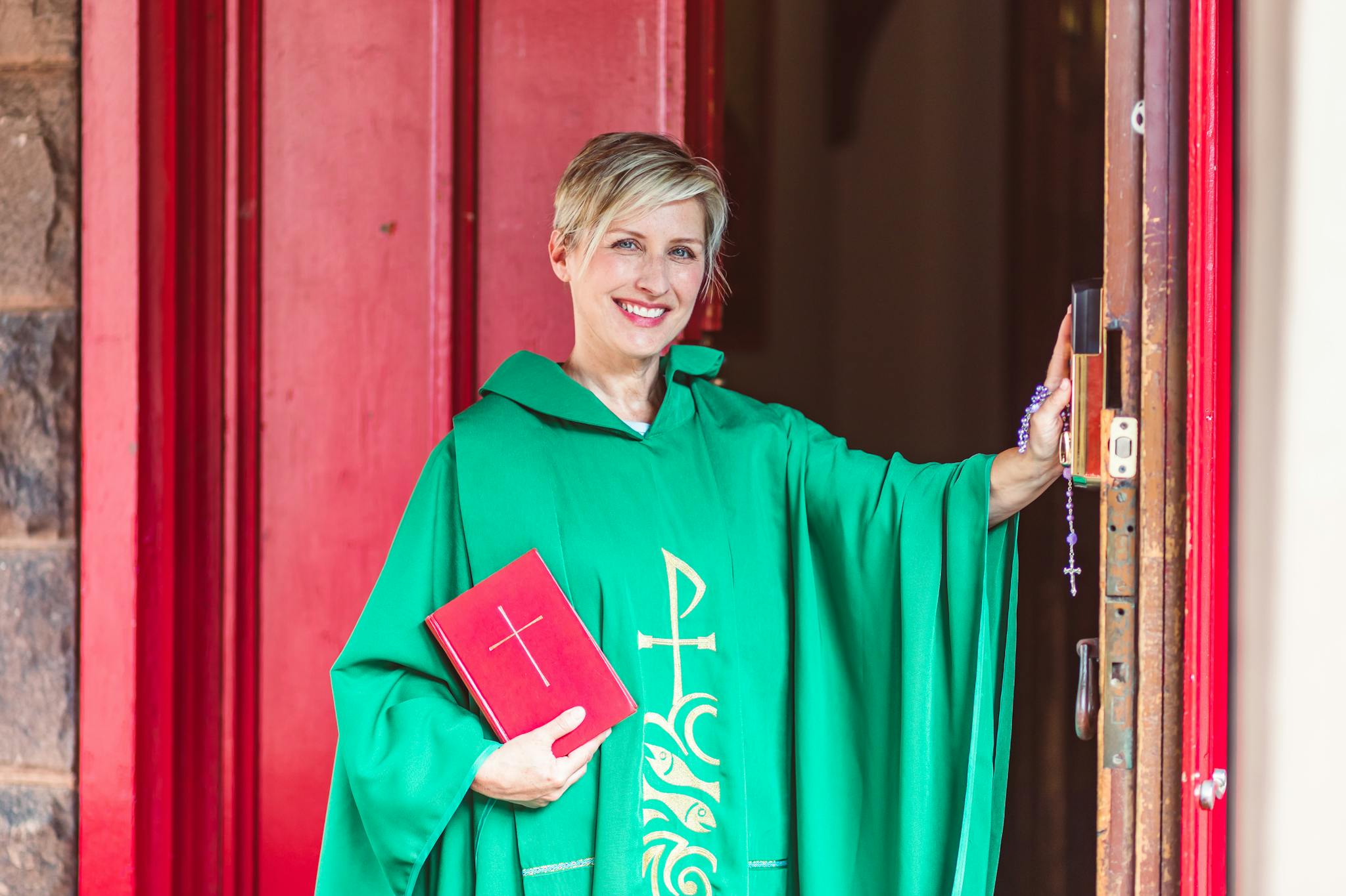 A smiling priest in a green cassock holding a Bible at a church doorway.