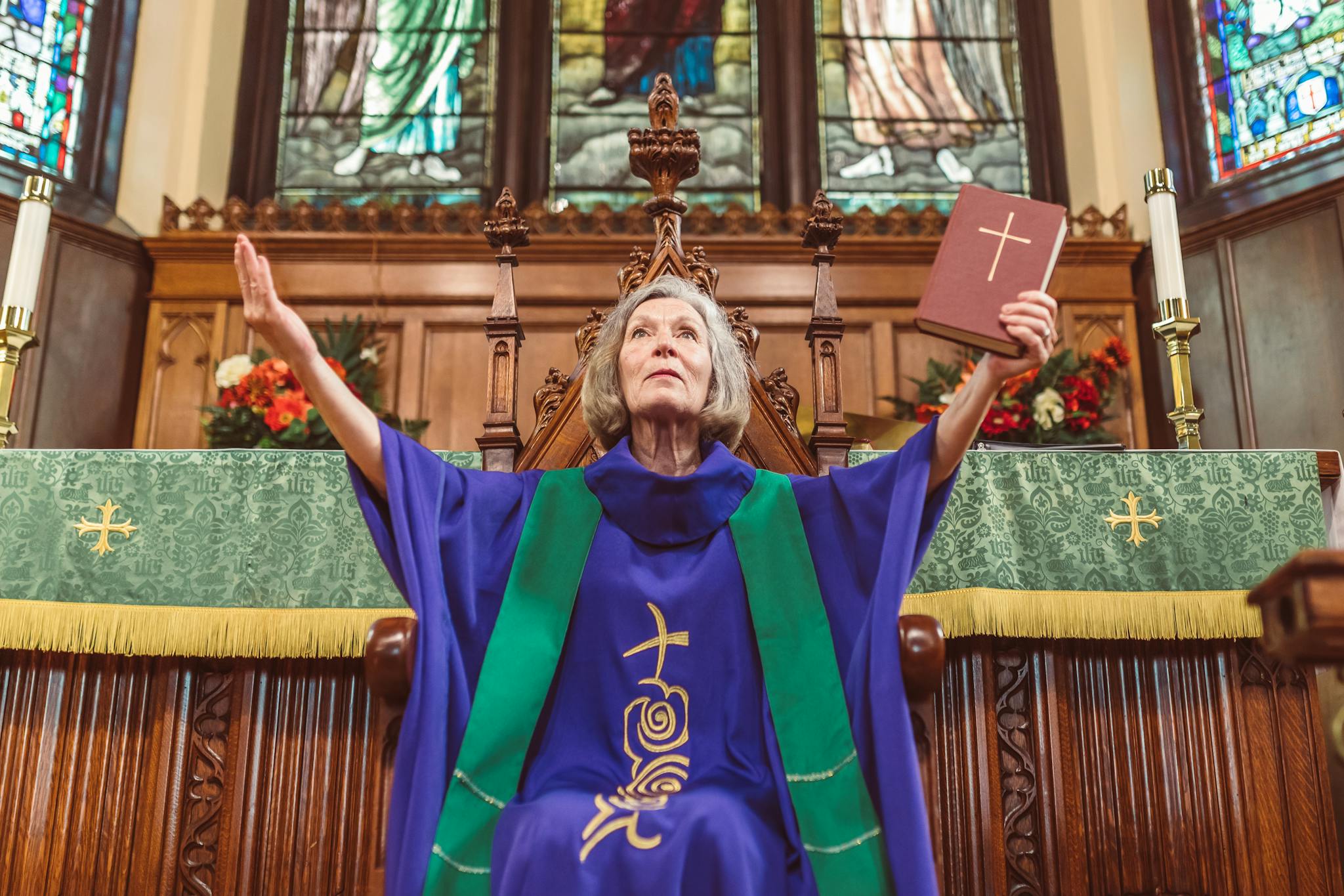 Elderly clergy woman in robes praising at church altar, holding a Bible.